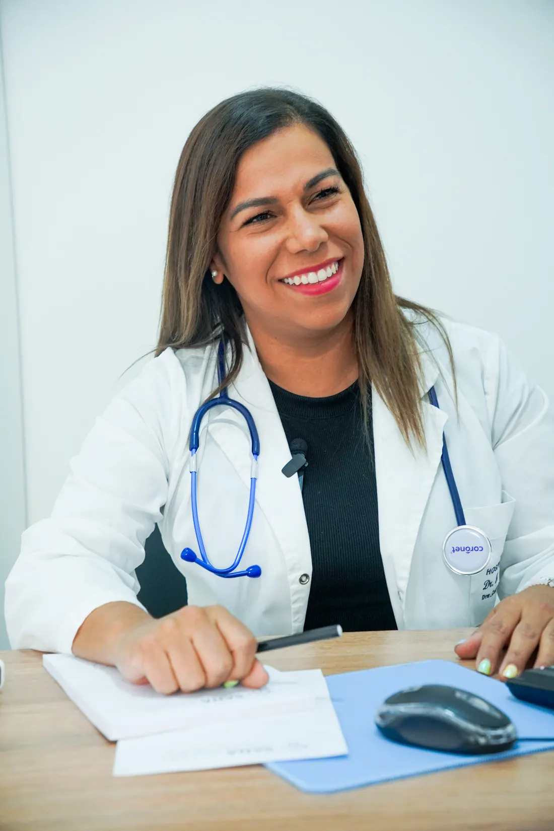 Médica de Sana Salud sonriendo en su consultorio, con estetoscopio, durante la atención de pacientes.